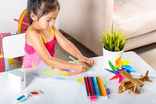 Happy child little girl colorful drawing landscape my home dream on paper, Asian cute kid preschooler sit on table smile she draw country house picture with pencil at home to learning arts homework