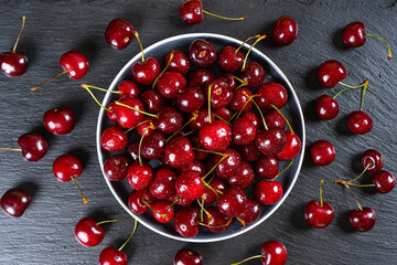Fresh red ripe sweet cherry on plate and scattered berries on black slate, stone background. Berry, food background. Top view, flat lay.
