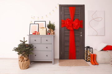 Beautiful fir tree near chest of drawers and wooden door decorated with red bow in room. Christmas decoration