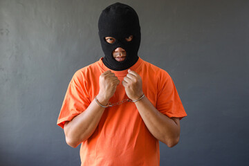 Portrait of prisoner in black mask and orange t-shirt wearing handcuff isolated over grey background