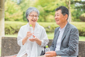 公園を散歩中に水を飲んで休憩するシニア・高齢者夫婦
