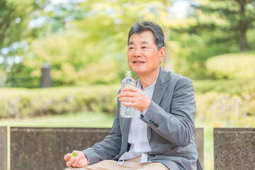 公園のベンチで水を飲む・水分補給するシニア男性
