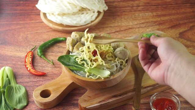 Meatball, in Indonesia known as Bakso or Baso. Served with noodles vegetables chili sauce in a bowl on wood background with hand. Close up top view flat lay copy space