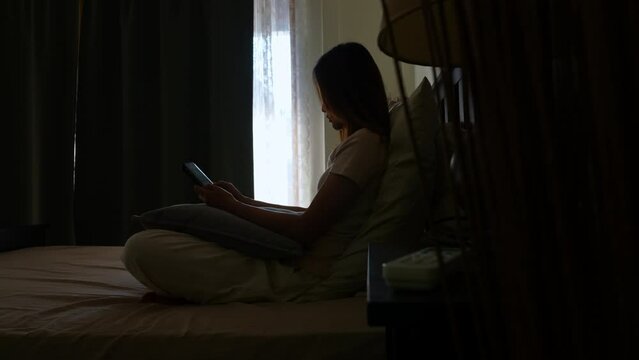 Depressed young Asian woman sitting on the bed and using mobile phone reading online message. Loneliness sad girl living alone with negative emotion and grief. Mental health and life problem concept.