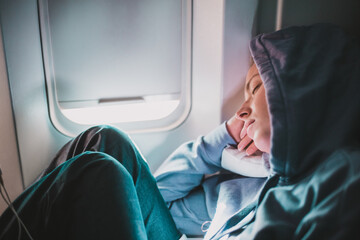 Tired blonde casual caucasian woman wearing sporty hoodie napping on seat while traveling by airplane. Commercial transportation by planes. Authentic image of real people - obrazy, fototapety, plakaty