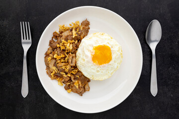 Khao Moo Tod Kratiam Kai Dao, Thai food, streamed rice topped with fried pork with garlic and fried egg in ceramic plate beside fork and spoon on dark tone texture background