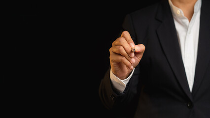 Businessman in a suit is holding a pen and writing in the air while standing on a black background