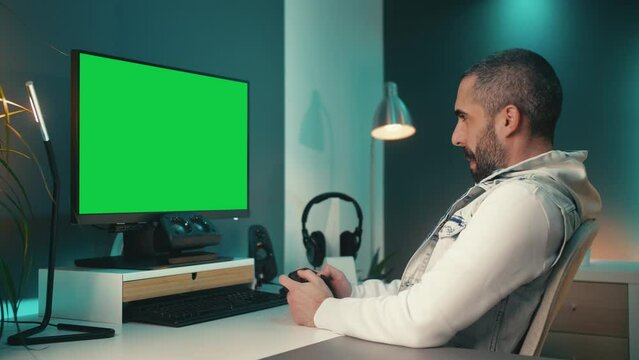 Young man plays a cool game on computer in colorful gaming studio. He has a green screen monitor and LED lights. He is immersed and excited by the game&rsquo;s graphics and sound effects.