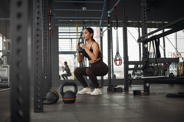 Strong Asian woman doing exercise with kettlebell at cross fit gym. Athlete female wearing sportswear doing squat workout on grey gym background with weight and dumbbell equipment. Healthy lifestyle.
