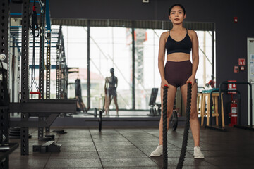 Strong Asian woman doing exercise with battle rope at cross-fit gym. Athlete female wearing sportswear workout on grey gym background with weight and dumbbell equipment. Healthy lifestyle.