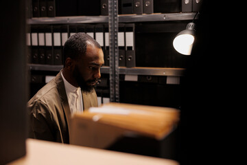 Concentrated african american cop working in police archive at night time. Man detective standing near shelves full of folders with records while conducting crime overtime