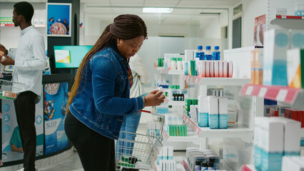 African american girl checking pharmaceutical products from pharmacy shelves, looking to buy medicaments and vitamins for healthcare. Young woman taking medicine and pills from drugstore.