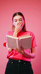 Surprising reading. Shocked literature. Astonished excited girl holding book covering mouth scared with storyline novel isolated on neon color pink background.