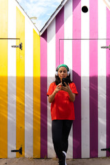 Beautiful latin woman leaning against a brightly colored striped booth using her mobile phone and music headphones on a bright summer day.