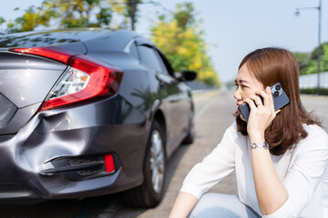 Upset unhappy Asian woman have a car accident or crashed during her road trip, woman driver sitting beside the crashed or broken car and using cell phone calling to road side assistant service.