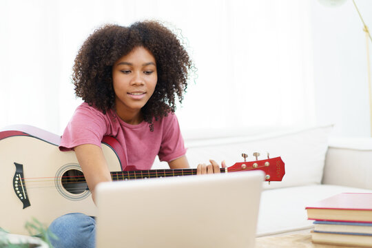 Happy cheerful American - African black woman sitting on a sofa in living room and playing an acoustic guitar close up with copy space.