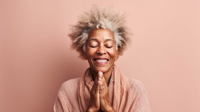 An afro senior woman smiles gently, her hands clasped in prayer in a light pink studio background. Generative AI