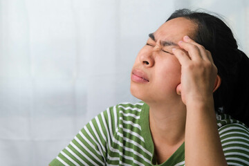 Young woman having headache against white curtain background in room. An adult woman touching head because of headache or migraine. Hands on head.