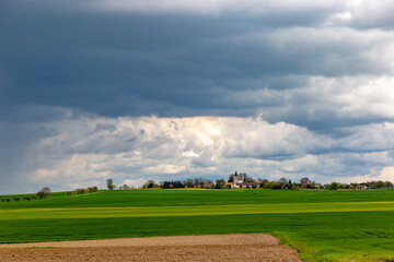 Fototapeta premium A small European village with a church on a hill, among spring fields, under a dramatic cloudy sky.