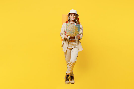 Full body cheerful happy young woman carry bag with stuff mat reading map isolated on plain yellow background. Tourist leads active lifestyle walk on spare time. Hiking trek rest travel trip concept.