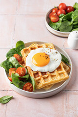 Savory breakfast with belgian waffles, fried egg and spinach, cherry tomato salad on pink tile background