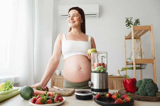 Smiling pregnant woman preparing fruit smoothie in kitchen at home