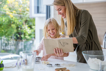 Happy woman reading recipe book by daughter preparing food in kitchen at home