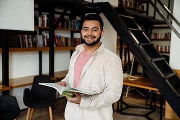 Cheerful indian man holding textbook while standing in library