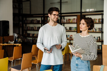 Man and woman students holding books and talking while standing in library