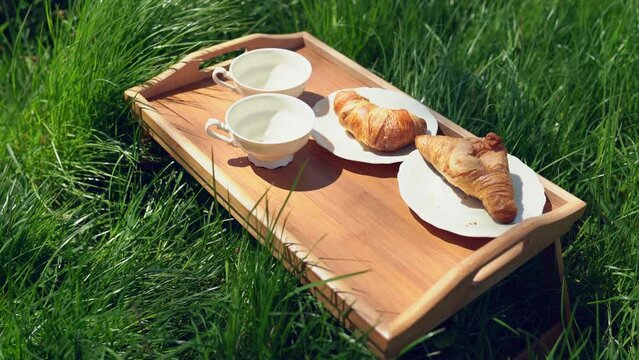 Porcelain coffee cups and plates with croissants on outside garden wooden tray. Sunny outdoor meal on green grass - romantic breakfast and picnic for two. Ruddy freshly baked croissant. 4k video 25FPS