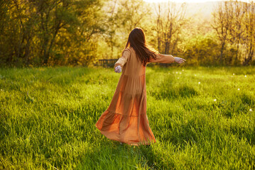 a relaxed slender woman enjoys the sunset standing in a green field with tall grass in an orange dress with her back to the camera, in warm summer weather. Horizontal photo