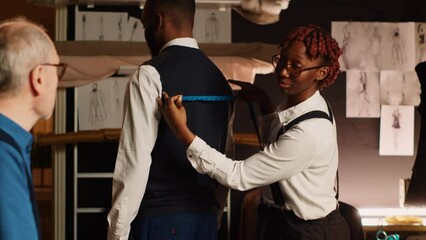Tailors team taking measurements for client, working in atelier to create custom made tailored suit. Couturier and assistant measuring customer to design perfect fit clothes. Handheld shot.