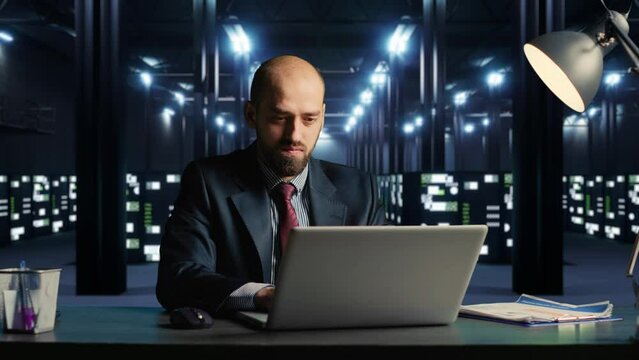IT engineer in suit working on laptop, sitting at desk in modern server room with cloud computing hardware. Businessman in data center with networking and security information. 3d render animation.