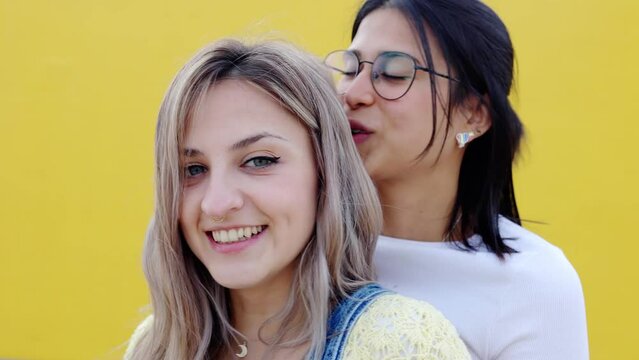 Portrait of smiling young lesbian couple hugging each other standing together over yellow background. LGBTQ people concept.