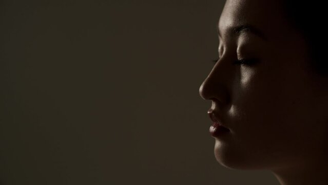 A young woman stands in a dimly lit room with her eyes closed. Beautiful Asian woman posing on a brown background.