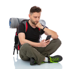 Portrait of serious trekker siting on the floor with a backpack looking for a tourist route on a smartphone map, isolated on white background. Thirty years old man posing in studio.