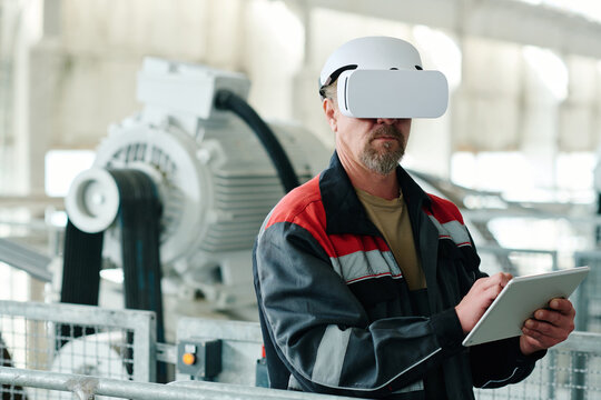 Mature engineer in uniform using digital tablet to watch production of material with VR glasses