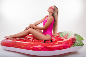 A beautiful blonde woman in a pink swimsuit and glasses is sitting on an air mattress on a white...