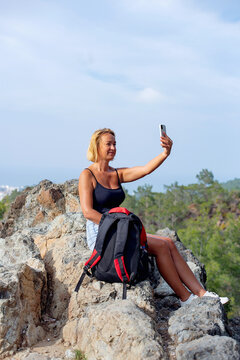 Woman traveler with a rucksack on back searching information on mobile smart phone while relaxing , young stylish female watching photos on cell telephone while sitting on a rock