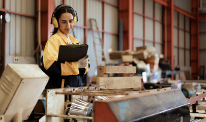 Female industrial engineer in white helmet, safety jacket and headphone work in heavy metal engineering factory. Latin technician woman worker using clipboard in metalwork manufacturing facility.