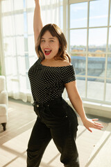 Obraz premium Emotional excited girl singing loudly with passion standing against big window putting hand up, looking at camera, wearing black jeans, dotted top and red lipstick, feeling herself as super star