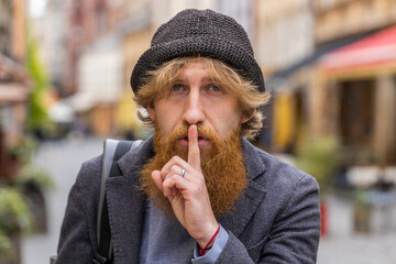 Shh be quiet please. Young bearded man presses index finger to lips makes silence hush gesture sign...