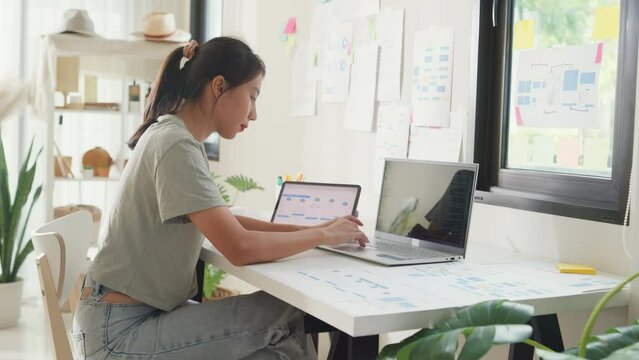 Young Asia girl IT development programmer typing on keyboard coding programming fixing data code on laptop computer screen follow code plan in tablet and paperwork on table workroom at house office.