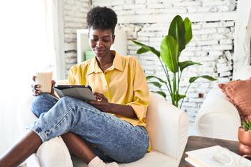 African woman enjoys breakfast and a video on her tablet, sipping milk in her living room.
