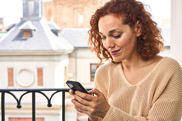 Redhead woman types on phone at balcony, immersed in urban view behind her.