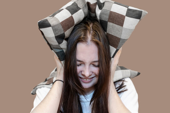 A young woman covers her head and ears with a pillow. The woman is upset, nervous and frowning.