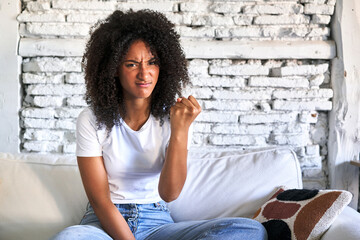Fototapeta na wymiar A young african american woman sitting on her sofa showing fist to camera, aggressive facial expression.