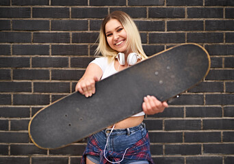 Happy young millennial student girl standing in front of a black brick wall and and looking at the camera, while showing a skateboard to the camera - People lifestyle concept with teenager © PEOPLEVISUALS