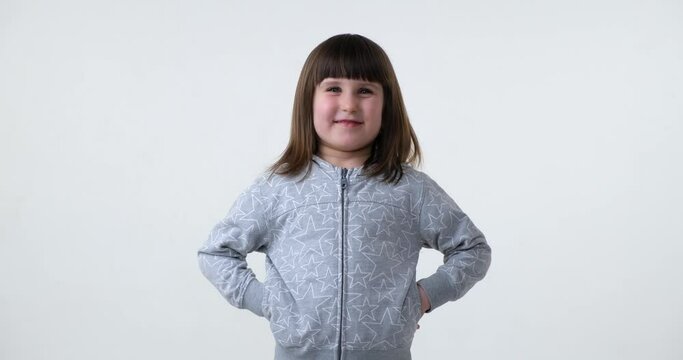A cute preschool-aged girl stands on a plain white background, looking straight into the camera with a bright smile on her face. She is dressed in casual clothing and her hair is styled neatly.