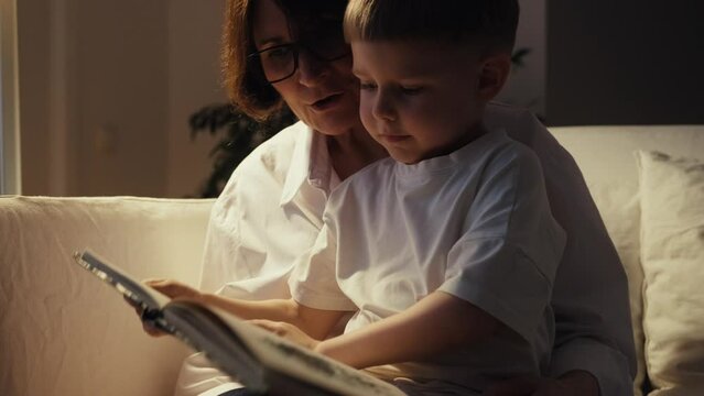 Grandmother and grandson are sitting on a white sofa and reading a book together. A cozy evening at Grandma's. Senior lady and her grandson spend time together.